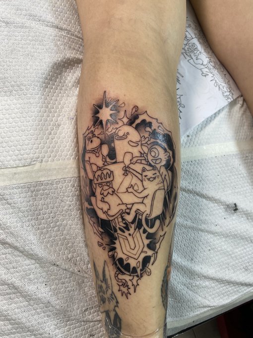 A black ink tattoo on someone's calf features cartoonish, grotesque characters including cats, a star, and a crown design, photographed against a white textured towel.