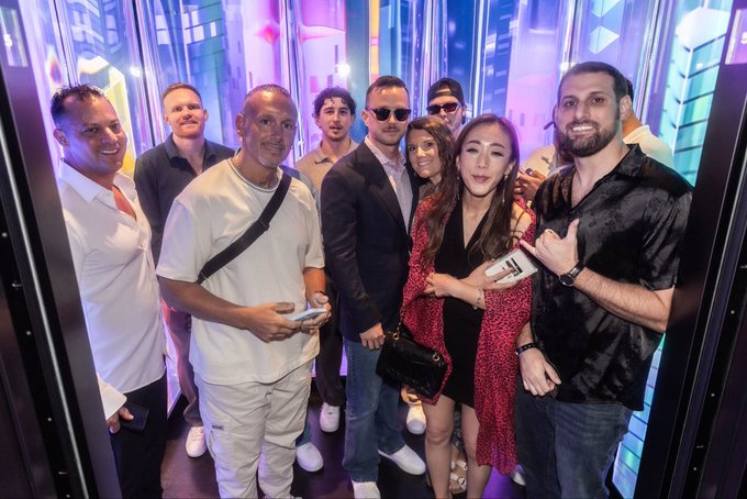 A group of seven well-dressed people posing together and smiling inside a brightly lit elevator with reflective walls.