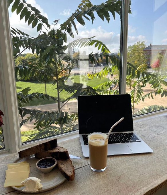 A laptop and an iced coffee sit on a wooden windowsill next to a plate of bread, cheese, and chocolate spread, overlooking a sunny view of a river and trees.