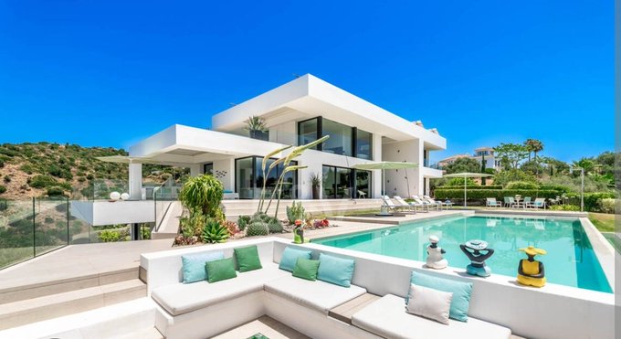 A modern white luxury villa with a swimming pool and outdoor lounge area featuring colorful pillows is set against a hillside under a clear blue sky.