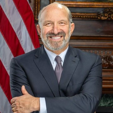 A man in a dark suit and patterned tie smiles with arms crossed, posed in front of an American flag. The post references Tom Brown of Anthropic, though this appears to be a different individual based on the image.

Without being able to confirm the identity of the person shown, I can't verify whether this image actually depicts Tom Brown or someone else. The image shows a smiling, bearded man in formal business attire in what looks like an official government setting, which could potentially be Howard Lutnick given the context of the post.
