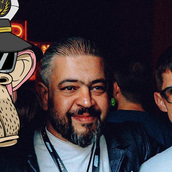 A bearded man with slicked-back hair smiles at the camera while wearing a lanyard at what appears to be an event in Lisbon, with an animated cartoon captain character edited into the left side of the frame, likely referencing the playful post about someone who "finally" looks like they're in their 20s rather than 13.