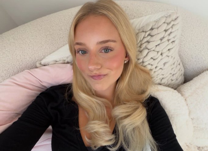 A selfie of a blonde woman with blue eyes, rosy cheeks, and pink lipstick, wearing a black top while sitting against white textured pillows.