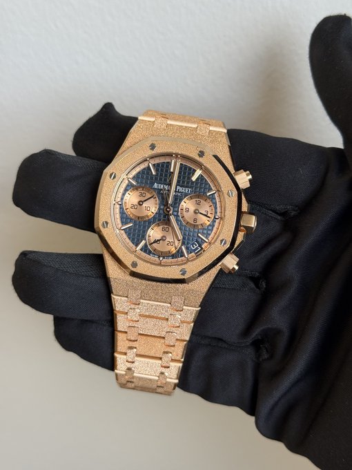 A gloved hand holds a gold-tone Audemars Piguet Royal Oak chronograph watch with a blue "Grande Tapisserie" dial, featuring sub-dials marked 30/10, 60, and 12/9, and a date window at 4:30. The luxury timepiece pairs with the post's greeting to crypto market participants, evoking themes of wealth, status, and the high-stakes trading world.