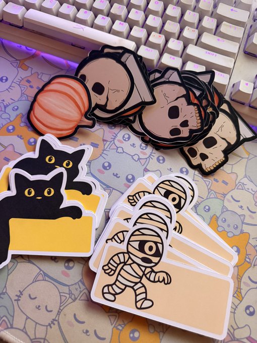 An assortment of cute Halloween stickers including black cats, mummies, skulls, and a pumpkin arranged on a pastel cat-themed desk mat next to a white keyboard.