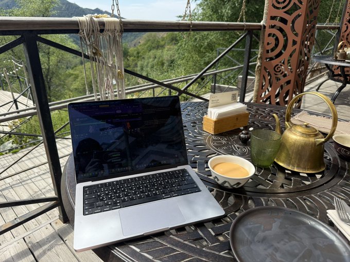 A laptop displaying trading charts sits on an ornate outdoor table alongside a brass teapot and coffee cup at Shymbulak mountain resort, capturing a serene work-from-anywhere moment while monitoring $MON perps live on Monad.