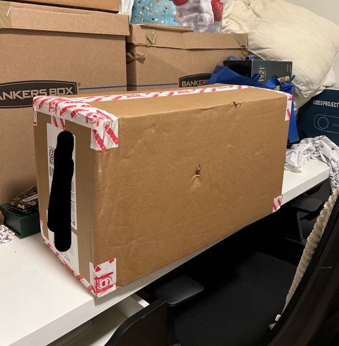 A cardboard shipping box secured with red and white packing tape sits on a desk alongside Bankers Box storage boxes, awaiting its unboxing. The package is poised to be opened by the traveler returning home, ready to reveal their long-awaited Azuki TCG packs in hopes of pulling a portrait card.