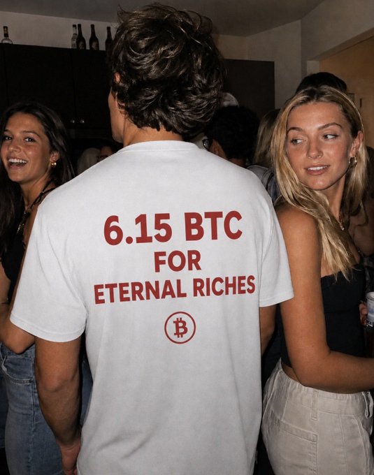 A young man wearing a white t-shirt reading "6.15 BTC FOR ETERNAL RICHES" with a Bitcoin logo stands in a social setting flanked by two women. The "Legend." caption fits the scene as the bold shirt declaration embodies the confident, meme-worthy swagger often celebrated in crypto culture.