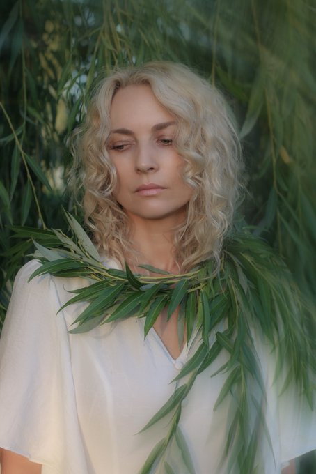 A portrait of a woman with curly blonde hair wearing a white top, standing among green willow leaves with her eyes closed.