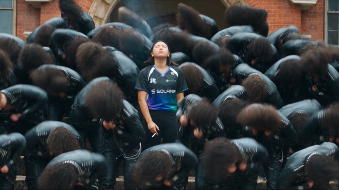 A cinematic image shows a person in a dark 'SOLANA' esports jersey standing in the center of a crowd of people bent over in dark clothing, with smoke rising in the background.