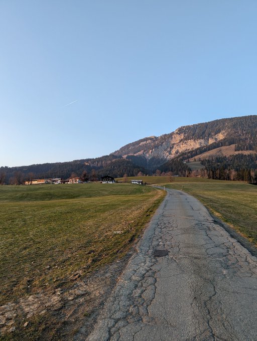 A scenic landscape photo of a cracked asphalt road winding through grassy fields toward a forested mountain under a clear blue sky with a visible contrail.
