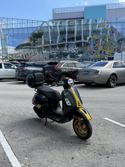 A dark green Vespa scooter with yellow racing stripes is parked in a parking lot, with several luxury cars visible in the background, suggesting the owner humorously swapped high-end status symbols for a modest ride.