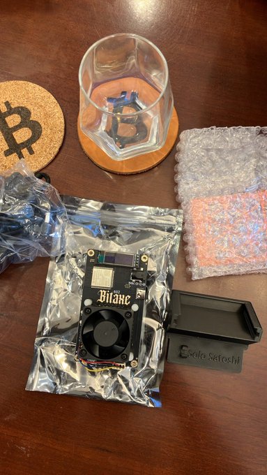 A Bitcoin hardware wallet setup on a wooden table, including a Bitaxe device in anti-static bag, a SafeSchiel enclosure, bubble wrap, a Bitcoin-themed coaster, and a glass with an etched Bitcoin logo.
