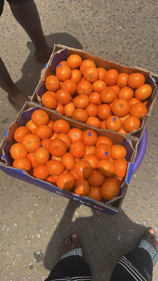 Two cardboard boxes filled with fresh oranges sit on a purple container on a paved ground, with a person's bare feet and patterned pants visible at the bottom and another person's leg on the left.