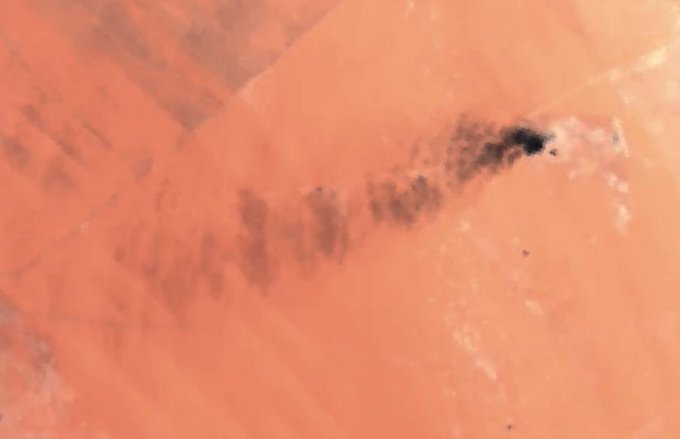 An aerial view showing dark smoke rising and spreading over a vast orange desert landscape.