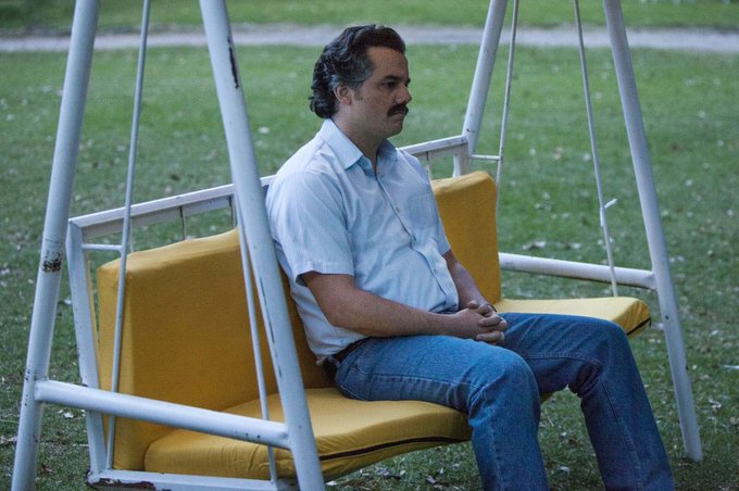 A man with a mustache wearing a light blue shirt and jeans sits alone on a yellow cushioned porch swing in a grassy yard.