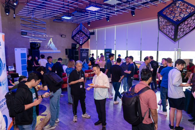 The image shows a large group of people gathered at an event with a focus on artificial intelligence (AI) and blockchain technology. The attendees are standing around, engaged in conversations and enjoying food from various stalls set up throughout the venue. Some individuals have backpacks or handbags, indicating that they might be carrying their belongings while attending the event.

The event appears to be taking place at a convention center with a spacious room filled with people, as well as several booths showcasing different aspects of AI and blockchain technology. The atmosphere seems lively and social, with attendees mingling and sharing ideas in this tech-focused gathering.