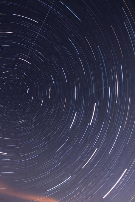 A long-exposure astrophotography shot capturing concentric circular star trails against a dark night sky, with a faint horizon glow at the bottom edge.