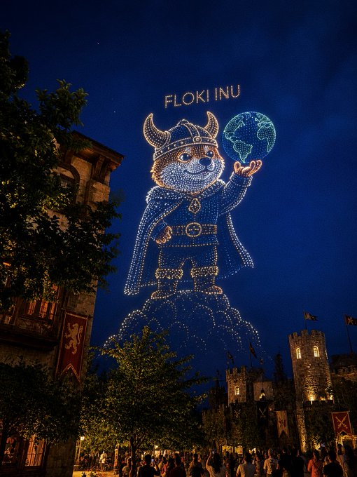 A night scene showing a large illuminated drone display of a Viking-like character with 'FLOKI INU' text and a globe, set against a castle-like building.