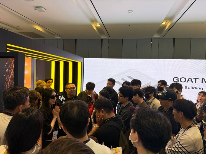 A crowded indoor gathering takes place in a modern venue with yellow neon accents, where attendees are gathered in front of a screen displaying 'GOAT M... Building P'.