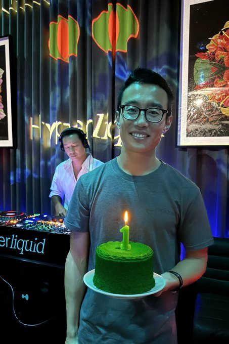 A smiling man in glasses holds a small green cake with a single candle shaped like the number "1," celebrating at what appears to be a Hyperliquid event (with the "Hyperliquid" logo visible behind him) while a DJ performs in the background—capturing the celebratory mood of $HYPE reaching an all-time high.