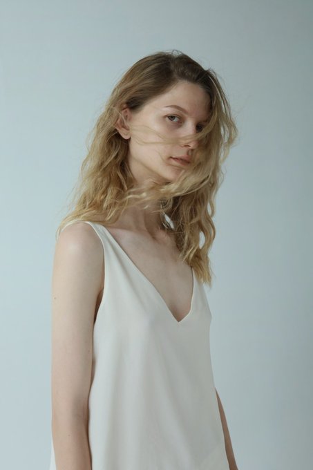 A portrait of a young woman with long, wavy blonde hair wearing a white sleeveless v-neck top, gazing softly at the camera against a plain light blue-gray background.