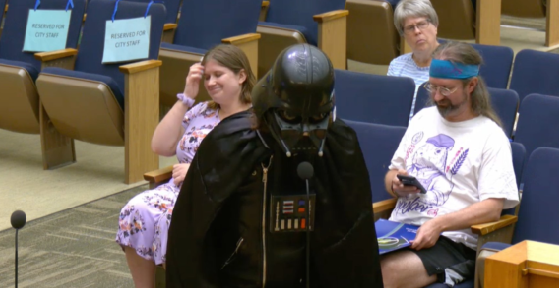 A person in a full Darth Vader costume stands at a podium in what appears to be a government chamber, while attendees including a smiling woman in a floral dress and a man in a graphic t-shirt with a headband look on from reserved seats marked "RESERVED FOR CITY STAFF." The post jokingly frames the Star Wars villain as a frustrated crypto trader pleading with attendees to stop short-selling his position at the town hall.