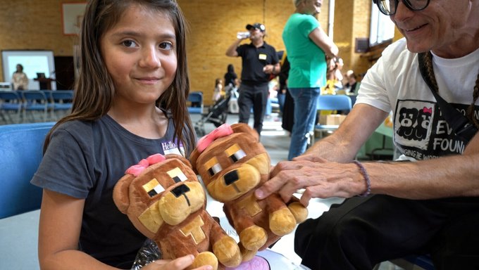 A young girl smiling while holding two brown teddy bear plush toys, with an older woman wearing glasses reaching toward the bears in what appears to be a community center.
