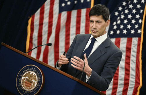 A man in a dark suit stands at a podium gesturing while speaking, with the U.S. flag behind him and the Department of State seal visible on the podium. The image relates to the post as a visual reference to Fed commentary or policy signaling, with the speaker representing the kind of official remarks—like Jackson Hole addresses—that shifted September rate hike probabilities and forced markets to reprice liquidity conditions across equities, metals, and crypto.