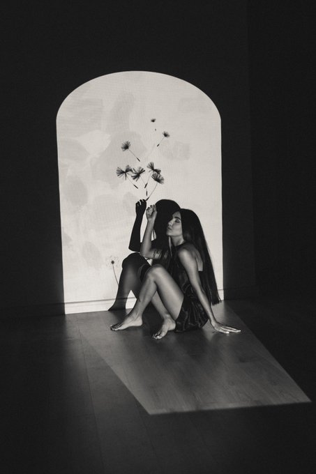 A black-and-white artistic photograph shows a woman sitting on the floor holding flowers, with light streaming through an arched doorway illuminating her and casting a window-pane shadow.