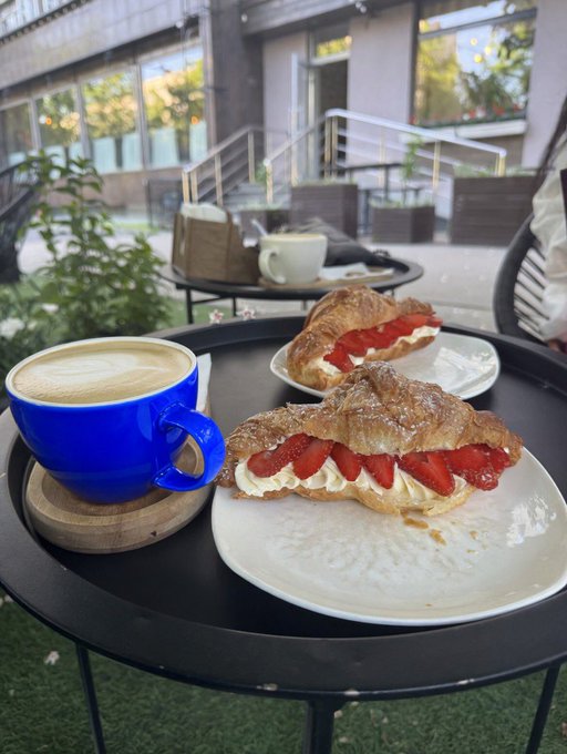 An outdoor café scene featuring a blue cup of cappuccino and a croissant filled with strawberries and cream on a plate atop a black table.
