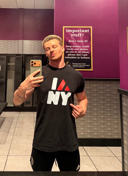 A mirror selfie of a man with blonde hair wearing an 'I ❤ NY' t-shirt, standing in front of a sign reading 'important stuff! don't lose it!' in a locker area.
