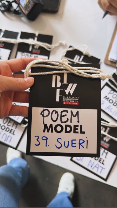 A hand holds up a black name tag on a string labeled "BIWF 1 (SIAM PARAGON, Bangkok Fashion Week, Fashion Week 2025) POEM MODEL 39. SUERI," with several similar tags scattered on a table below, suggesting behind-the-scenes credentialing at Bangkok Fashion Week.