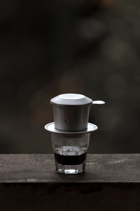 A Vietnamese coffee filter (phin) sits on top of a glass containing dark coffee, photographed on a ledge with a blurred background.
