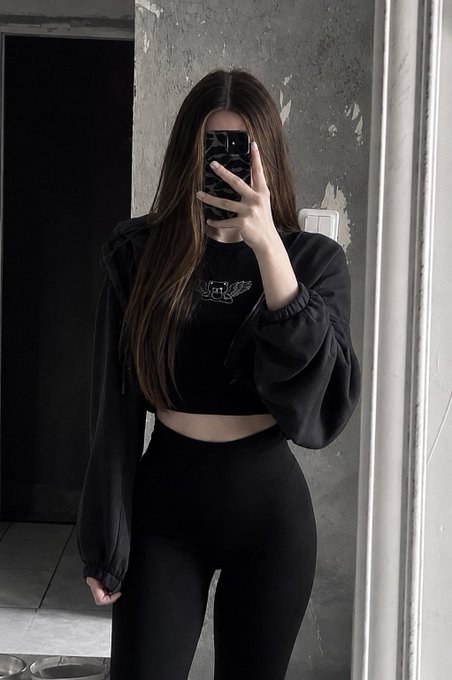 A person takes a selfie with a black phone, wearing a black crop top and leggings against a gray wall backdrop.