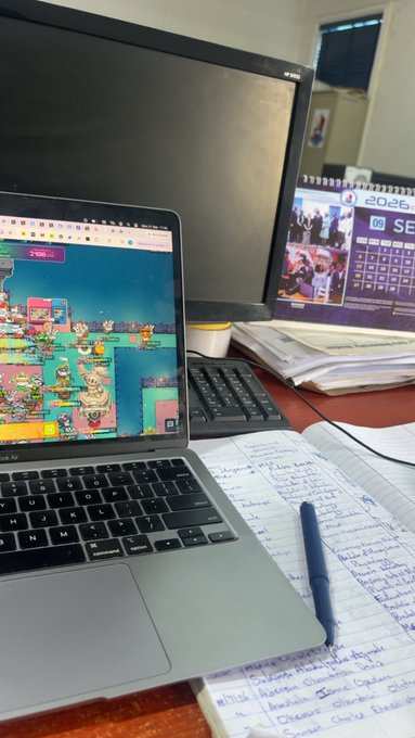 A photograph of a desk workspace showing an open laptop displaying colorful cartoon images, next to a notebook filled with handwritten notes and a pen, with a calendar on the monitor in the background.