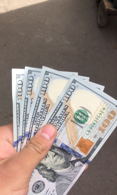 A hand holds a fan of several US $100 bills featuring Benjamin Franklin, photographed over a gray asphalt surface.