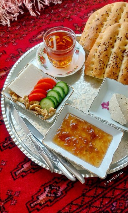 An overhead view of a Persian breakfast spread featuring flatbread (naan), white cheese, feta, walnuts, sliced tomatoes and cucumbers, honey, and tea, set on a silver tray atop a red patterned rug, with a watermark 'S DEHGHANI'.