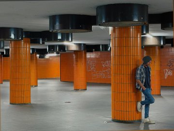 A lone figure in a plaid shirt and cap leans against an orange-tiled pillar in a graffiti-tagged underground hall, evoking the gritty, moody atmosphere of Berlin's U-Bahn stations—fitting the "Berlinmaxxing" theme of urban exploration and aesthetic immersion in the city.