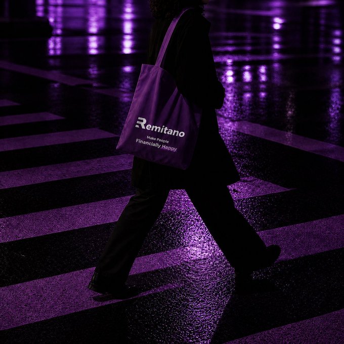 A stylish photograph of a person in black clothing walking across a crosswalk, carrying a purple tote bag with the 'Remitano' logo and tagline 'Make People Financially Happy.'