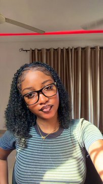 A selfie of a young woman with curly dark hair, glasses, and a blue and white striped shirt, taken indoors with red lighting visible in the background.