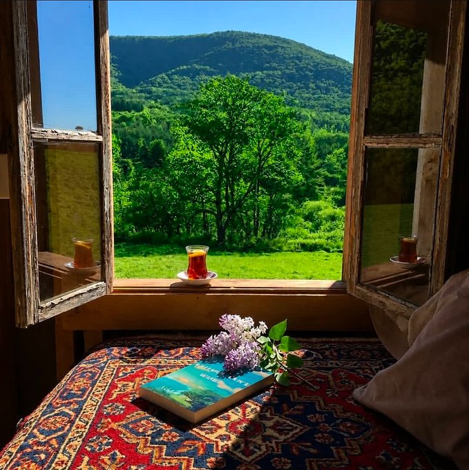An open wooden window frames a lush green mountain landscape, with a glass of tea resting on the windowsill and a book titled 'A View from the Bridge' with lilacs placed on a patterned rug in the foreground.