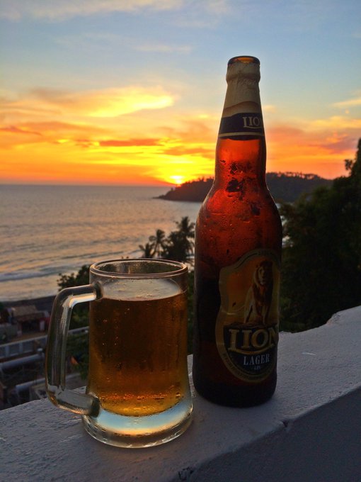 A bottle of Lion Lager beer and a glass mug filled with beer sit on a ledge overlooking a tropical ocean at sunset.