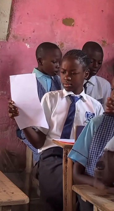 Three young school children in white shirts and blue ties stand at desks in a classroom with pink walls; the central child is reading from a piece of paper.