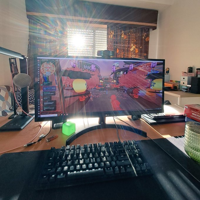 The image shows an office desk with a computer monitor displaying a video game scene, likely from the Nintendo Wii console. The monitor is turned on, and there are two keyboards placed next to each other on the desk. A mouse can also be seen in front of the keyboard area. In addition to these peripherals, there is a cup positioned near the edge of the desk. The image also contains text that reads "Had a little moment with @decentraland last evening, after a while".