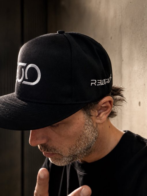 A man with stubble wearing a black 'R3GRT' baseball cap looks downward, with the cap's brim casting a shadow over his face against a concrete wall background.