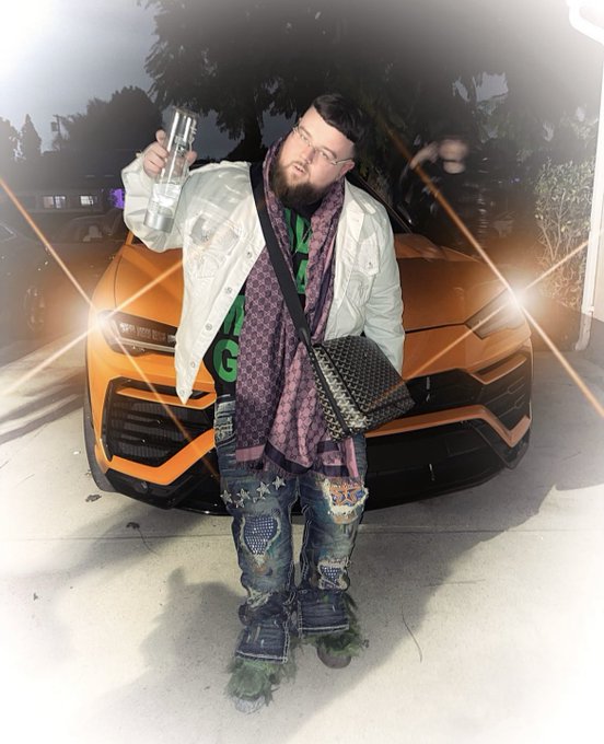 A man stands in front of an orange Lamborghini SUV at night, wearing a white jacket, green shirt, patterned scarf, and carrying a Louis Vuitton bag.