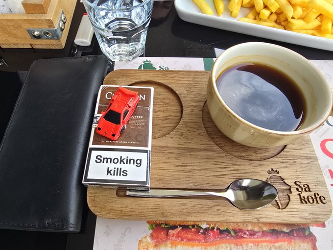 A coffee cup, a spoon, and a "Smoking kills" cigarette pack rest on a wooden tray alongside a wallet, with fries and a menu visible nearby.