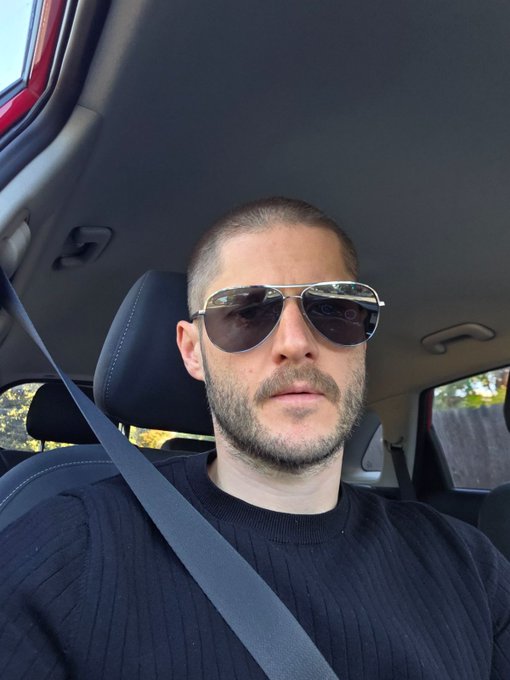 A bearded man wearing aviator sunglasses and a black sweater sits in the driver's seat of a car with his seatbelt on, projecting the laid-back, authentic "no longer trying to impress anyone" vibe of the accompanying post.