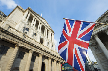 The Bank of England building in London stands prominently under a bright sky, with the Union Jack flag waving in the foreground, symbolizing the UK's financial authority where HM Treasury is proposing new "innovate, but don't destabilize" objectives to support innovation in payment systems and digital money, including stablecoins.